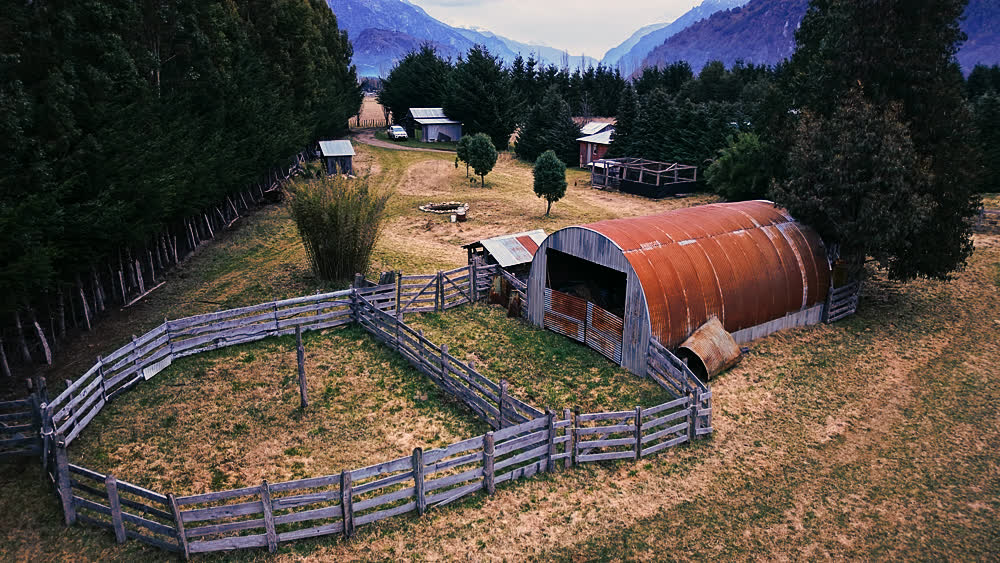 Vista aérea de un galpón de metal oxidado junto a corrales de madera, con la cordillera de fondo